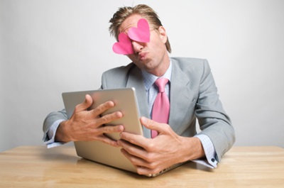 White man in a grey suit sits at a desk with his arms around an open laptop computer. He has pink paper heart shapes on his eyes.
