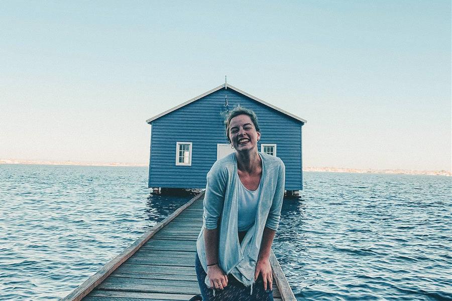 Young woman kneeling and smiling on jetty outside the blue boat shed in Perth