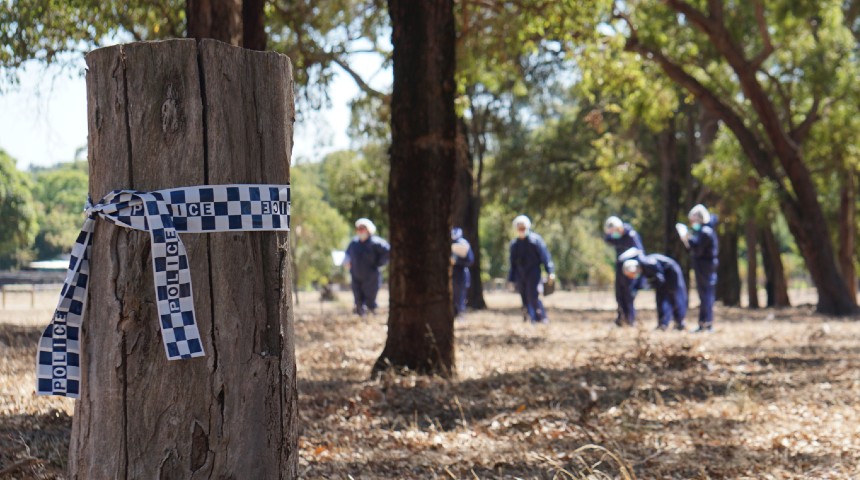 With police tape in foreground, a group of students in boiler suits walk though forest and investigate a mock-crime scene