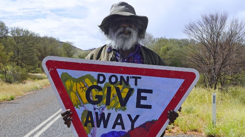 Iltja Ntjarra artist Mervyn Rubuntja holding the Don't Give Away hand-painted sign.