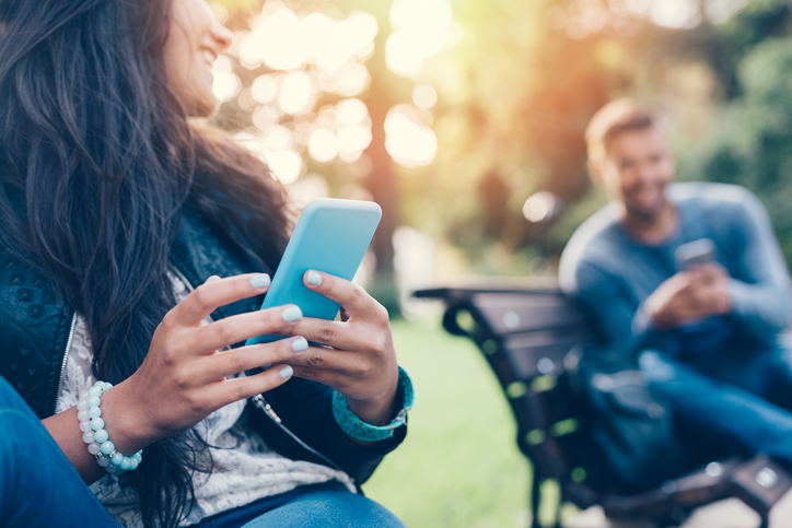 Woman and man sitting on park benches, smiling and looking at phones.