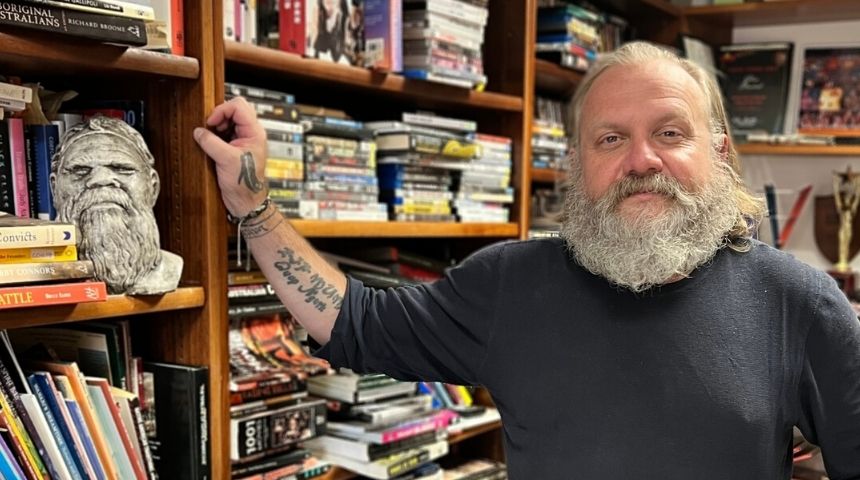 Dr Glen Stasiuk, a white male with a grey beard, in his office with shelves of books and dvds in the background