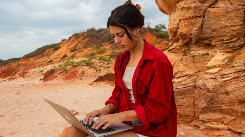 Student in red shirt sits on a red rock and studies laptop