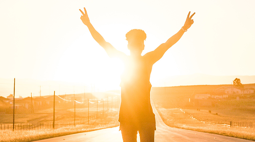 Boy standing on the road with his hands in the air and the sun setting behind him