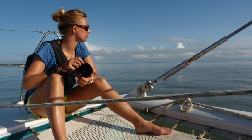 Blonde girl on left hand side of a sailing boat looking at the ocean and holding a camera.