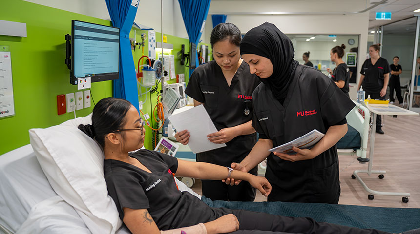 Two female students take the pulse of a third student who is lying on a bed in the SimLab.