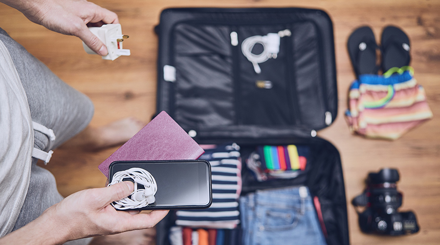 Man standing over suitcase filled with clothes holding a phone, passport, and adaptor