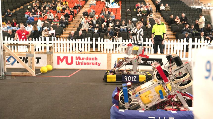 Two robots in the foreground in an indoor fenced area with stadium seats behind the short fence. Two people inside the fence with the robots and spectators in the stands.