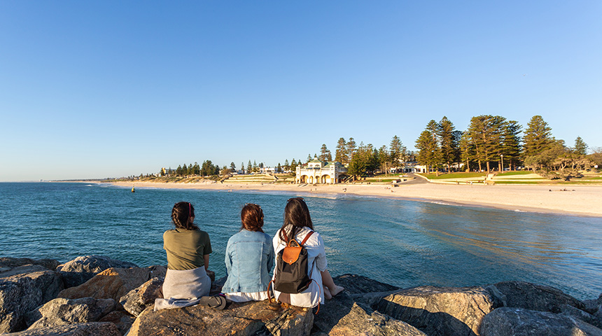 Three female students sitting with their backs to the camera on the groyne at Cottesloe beach with the Indiana Teahouse in the background.