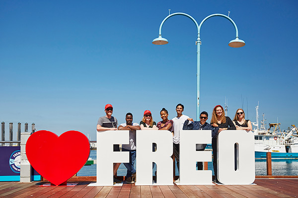 Group of smiling students standing behind a 'love freo' sign on habour in Fremantle 