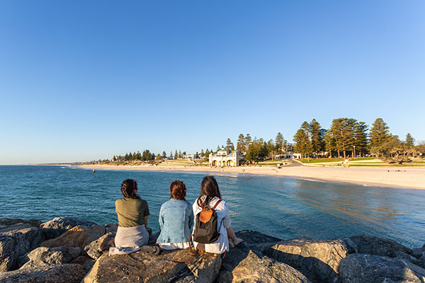 Three female students sitting with their backs to the camera on the groyne at Cottesloe beach with the Indiana Teahouse in the background.